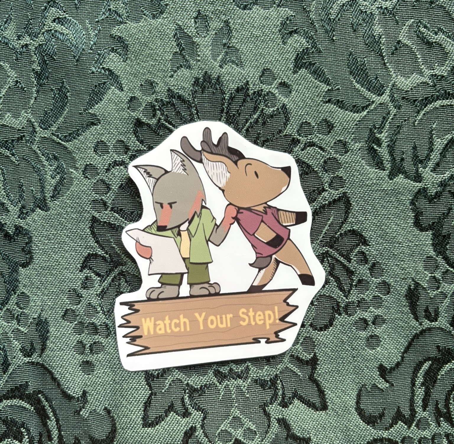 Picture of a coyote looking at a map with one hand. Their other hand stops a mule deer from walking off the side of a cliff. Text reads WATCH YOUR STEP.