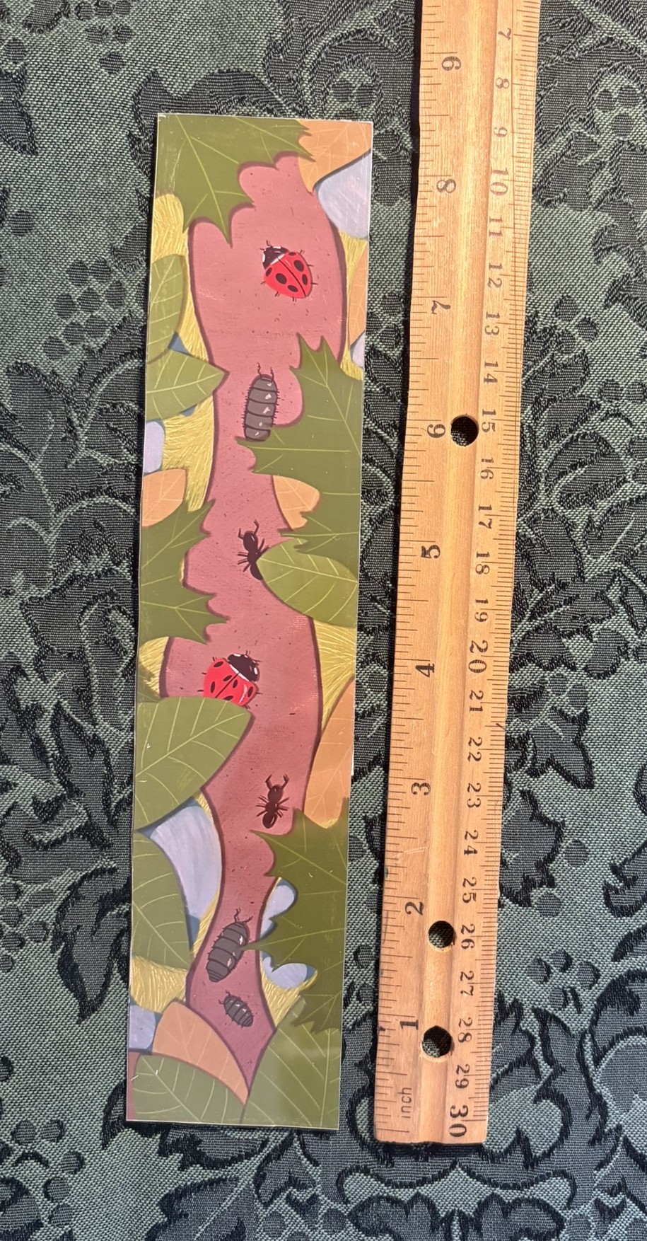 A glossy bookmark depicting a small forest trail covered by leaves. Ladybugs and pillbugs can be spotted on the trail.